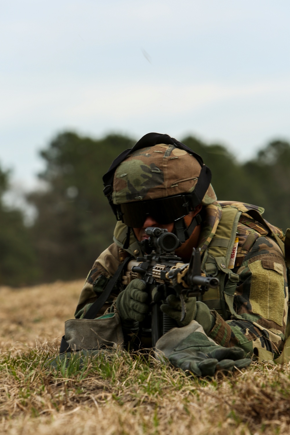 A soldier in full combat gear is in a prone firing position on open grassland, aiming a rifle with an optical sight. The individual is wearing woodland/multicam camouflage uniform, a Kevlar helmet with camouflage cover, tactical backpack, and protective eyewear. The background shows a treeline under an overcast sky.