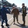 USNS Wheeler Performs Demonstration, Holds Tour for Military Officials Military personnel in various uniforms are gathered on what appears to be a naval pier or dock facility. A handshake greeting is taking place between individuals wearing U.S. Navy blue digital camouflage and U.S. military tan/coyote brown gear, with a Marine in woodland MARPAT camouflage standing nearby. The setting suggests an official meeting or arrival ceremony at a port installation.