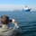 USNS Wheeler Performs Demonstration, Holds Tour for Military Officials A person in military camouflage uniform is photographing a large blue cargo or offshore supply vessel from a smaller watercraft. The ship in the background appears to be carrying heavy industrial equipment or machinery on its deck. The scene takes place on open ocean water with a coastline and hazy skyline visible in the distance.