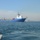 USNS Wheeler Performs Demonstration, Holds Tour for Military Officials A large blue and white offshore supply or cable-laying vessel is underway on calm open water. The ship's deck is loaded with large industrial spools or reels, likely containing subsea cable or pipeline. A port or industrial facility is visible in the hazy background on the right.