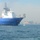 USNS Wheeler Performs Demonstration, Holds Tour for Military Officials A large blue and white offshore support or survey vessel is photographed at sea, with an industrial port or coastal facility visible in the background. The ship appears to be discharging water from its hull, visible as a jet stream near the bow. The vessel displays a yellow and blue stripe on its superstructure, consistent with a specialized offshore service company livery.