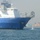 USNS Wheeler Performs Demonstration, Holds Tour for Military Officials The image shows a large blue and white U.S. Naval Ship named 'VADM K R WHEELER' anchored or underway in a harbor. A water jet or discharge stream is visible near the bow of the vessel, likely from a ballast or cooling water discharge port. An industrial port facility with shipping containers and structures is visible in the background.