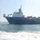 USNS Wheeler Performs Demonstration, Holds Tour for Military Officials A large blue-hulled offshore supply or cable-laying vessel is photographed from water level, likely from a smaller moving vessel given the wake visible in the foreground. The ship carries large cable reels or drum spools on its deck along with various industrial equipment and a white superstructure. The image is taken in open coastal waters with hazy sky conditions and distant hills visible on the horizon.