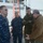 USNS Wheeler Performs Demonstration, Holds Tour for Military Officials Four military personnel are gathered dockside near a naval vessel, engaged in a discussion or briefing. Three individuals are wearing military uniforms — two in Navy Working Uniform (NWU) digital blue camouflage and one in Marine Corps MARPAT camouflage — while a fourth person wears a tan/coyote fleece jacket with Navy-style trousers. The setting appears to be a naval shipyard or pier facility.