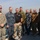 USNS Wheeler Performs Demonstration, Holds Tour for Military Officials A group of eight military personnel and one civilian are posing for a group photo on the deck of a vessel, identifiable by the word 'APPLETON' visible on a railing. The individuals are wearing a variety of military uniforms representing multiple branches and possibly multiple nations, including US Air Force, US Navy, US Marine Corps, and what appears to be a Canadian or allied military uniform. The photo is taken outdoors over open water with a hazy sky in the background.