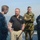 USNS Wheeler Performs Demonstration, Holds Tour for Military Officials Four individuals are gathered on the deck of a naval vessel near open water. Two are wearing U.S. Navy blue digital camouflage uniforms, one is wearing U.S. Marine Corps MARPAT woodland camouflage, and one is in civilian attire with a black polo shirt. They appear to be engaged in a briefing or discussion.