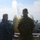 USNS Wheeler Performs Demonstration, Holds Tour for Military Officials Two military personnel are standing on the deck of a naval vessel observing a high-pressure water cannon or fire monitor in operation, projecting a powerful stream of water into the air toward the sea. One individual is wearing U.S. Navy blue digital camouflage (NWU Type I) and the other is wearing U.S. Marine Corps MARPAT woodland camouflage. The scene appears to be a demonstration or training exercise conducted at sea.