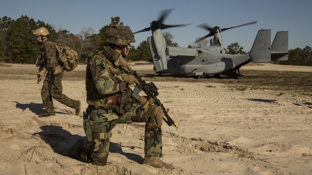 Military personnel are conducting an insertion or extraction operation with a Bell Boeing V-22 Osprey tiltrotor aircraft on a sandy landing zone. One soldier in woodland camouflage pattern is kneeling in a security position with a rifle while another in MARPAT digital camouflage moves toward the aircraft. The 'MARINES' marking is visible on the Osprey's tail, indicating U.S. Marine Corps involvement.