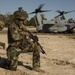 Military personnel are conducting an insertion or extraction operation with a Bell Boeing V-22 Osprey tiltrotor aircraft on a sandy landing zone. One soldier in woodland camouflage pattern is kneeling in a security position with a rifle while another in MARPAT digital camouflage moves toward the aircraft. The 'MARINES' marking is visible on the Osprey's tail, indicating U.S. Marine Corps involvement.