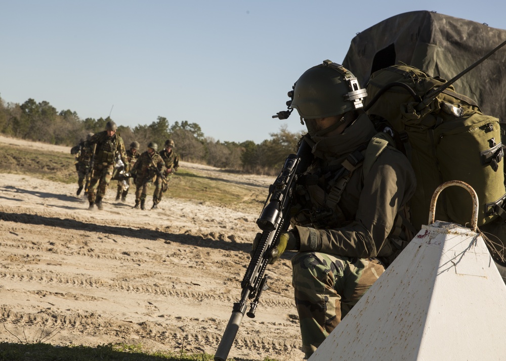 A military training exercise is depicted on a sandy dirt road surrounded by sparse woodland. A foreground soldier in full tactical gear including helmet with mounted device, large rucksack, and rifle takes a covered position behind a white barrier, while a group of approximately four soldiers in camouflage uniforms approach in the background. The setting and equipment suggest a field training exercise rather than active combat.
