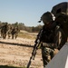 A military training exercise is depicted on a sandy dirt road surrounded by sparse woodland. A foreground soldier in full tactical gear including helmet with mounted device, large rucksack, and rifle takes a covered position behind a white barrier, while a group of approximately four soldiers in camouflage uniforms approach in the background. The setting and equipment suggest a field training exercise rather than active combat.