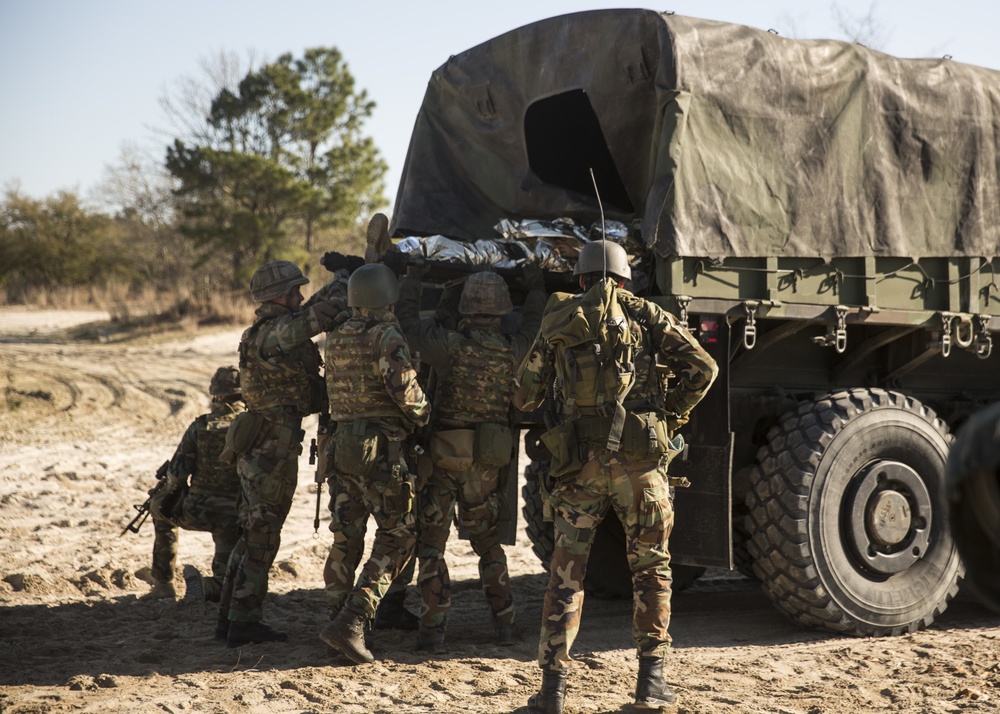 A group of military personnel in camouflage uniforms and helmets are gathered around the rear of a canvas-covered military cargo truck on a sandy dirt road. The soldiers appear to be loading or unloading cargo, possibly including a wrapped object visible in the truck bed. The setting appears to be a training area or field environment based on the terrain and sparse vegetation.