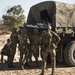 A group of military personnel in camouflage uniforms and helmets are gathered around the rear of a canvas-covered military cargo truck on a sandy dirt road. The soldiers appear to be loading or unloading cargo, possibly including a wrapped object visible in the truck bed. The setting appears to be a training area or field environment based on the terrain and sparse vegetation.