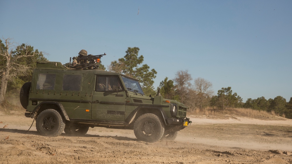 A military-configured Mercedes-Benz G-Class (Geländewagen) painted in olive drab is traversing a sandy dirt trail, kicking up dust. A soldier is positioned in a roof-mounted gun turret operating what appears to be a heavy machine gun. The setting appears to be a training area surrounded by pine and deciduous trees under clear blue skies.