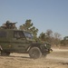 A military-configured Mercedes-Benz G-Class (Geländewagen) painted in olive drab is traversing a sandy dirt trail, kicking up dust. A soldier is positioned in a roof-mounted gun turret operating what appears to be a heavy machine gun. The setting appears to be a training area surrounded by pine and deciduous trees under clear blue skies.