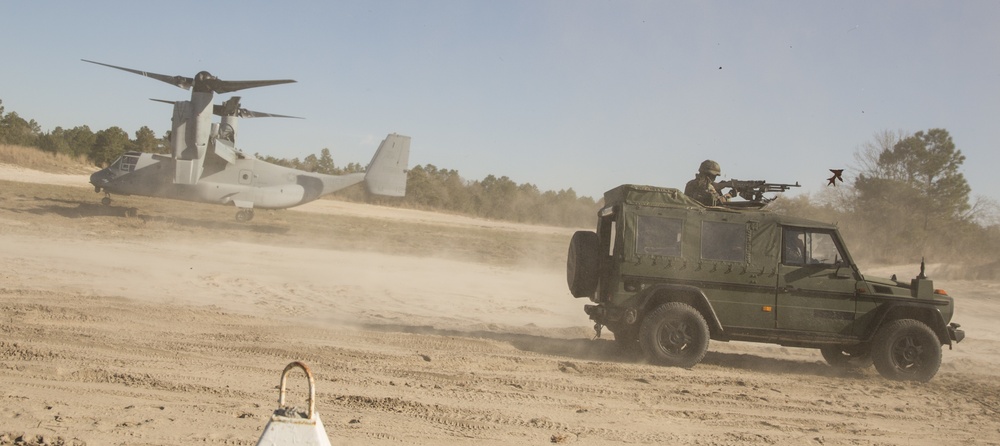 A military exercise or operation is underway on a sandy terrain, featuring a V-22 Osprey tiltrotor aircraft on the ground with rotors spinning and a military utility vehicle (Mercedes-Benz G-Class/Wolf variant) with a mounted machine gun and armed personnel. Dust is being kicked up by both the vehicle and the aircraft's rotor wash. The scene suggests a rapid insertion or extraction operation typical of Marine or special operations training.