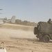 A military exercise or operation is underway on a sandy terrain, featuring a V-22 Osprey tiltrotor aircraft on the ground with rotors spinning and a military utility vehicle (Mercedes-Benz G-Class/Wolf variant) with a mounted machine gun and armed personnel. Dust is being kicked up by both the vehicle and the aircraft's rotor wash. The scene suggests a rapid insertion or extraction operation typical of Marine or special operations training.
