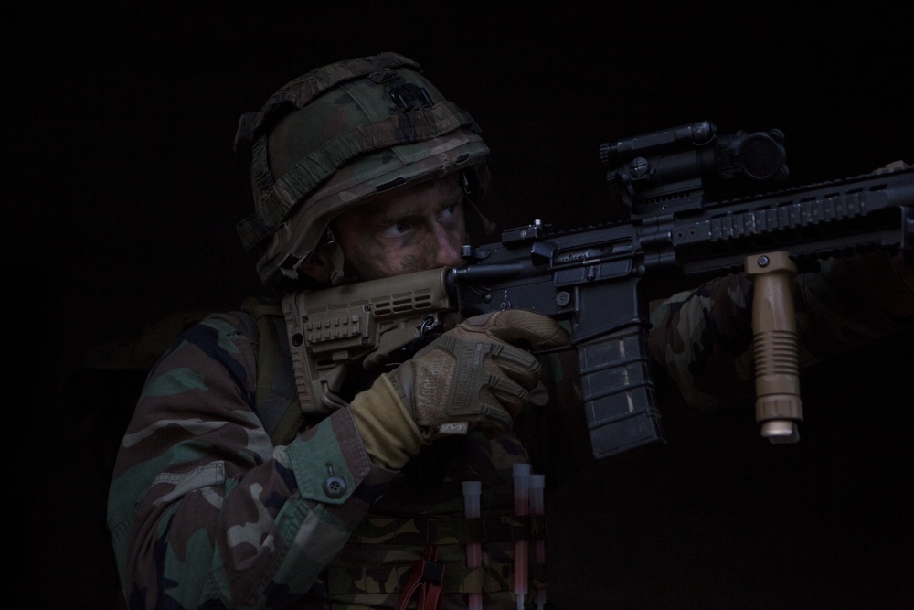 A soldier in full combat gear is shown aiming a rifle in a low-light environment. The individual is wearing camouflage uniform, a tactical helmet, and tan gloves while shouldering what appears to be an AR-platform rifle with multiple accessories. The dark background and dramatic lighting suggest a nighttime or indoor tactical scenario.