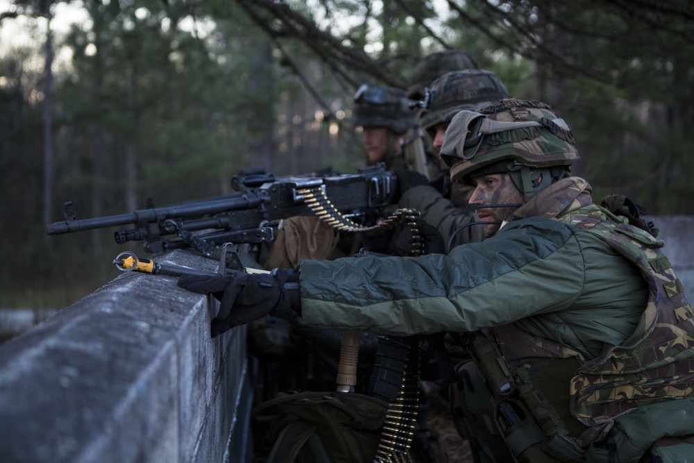 The image shows military personnel in full combat gear taking a defensive position behind a concrete barrier. They are operating what appears to be a belt-fed machine gun, with ammunition belts visibly draped across the weapon. The soldiers are wearing camouflage uniforms, helmets with covers, and face paint, suggesting a training exercise or tactical operation in a wooded environment.