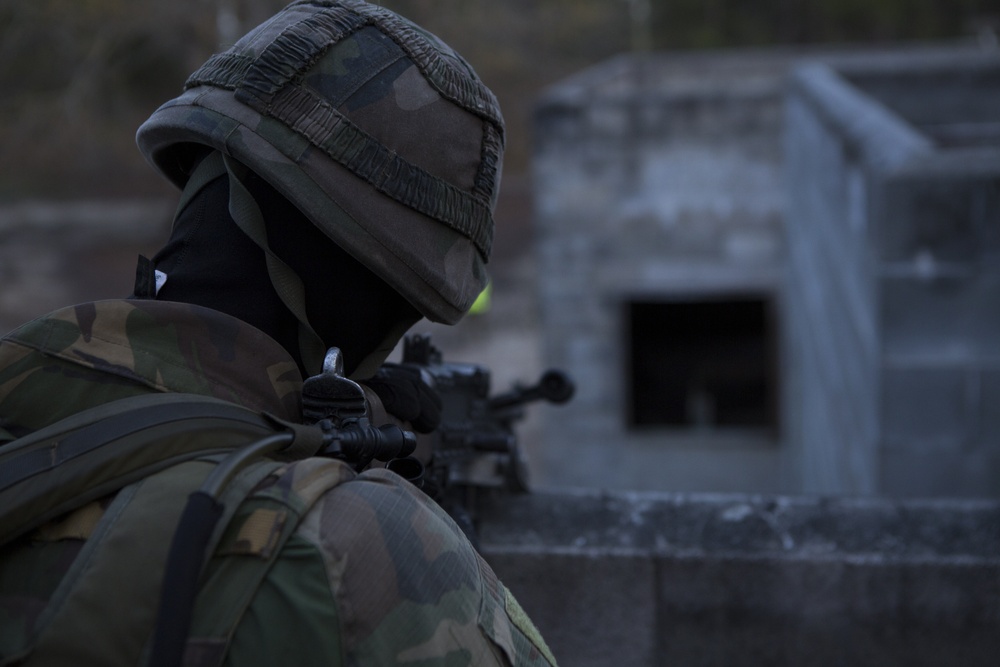 A soldier in camouflage uniform and helmet is positioned behind a low concrete wall, aiming a rifle toward a concrete structure in the background. The individual is wearing a black balaclava for face concealment and a camouflage-covered helmet. The setting appears to be a military training or operational environment with low ambient light.
