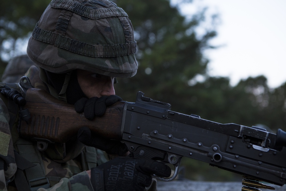 A soldier in camouflage uniform and helmet is aiming a machine gun, likely during a training exercise or field operation. The weapon appears to be a belt-fed machine gun, possibly an FN MAG or similar variant, with a visible ammunition belt. The image is taken at close range with a shallow depth of field, emphasizing the soldier and weapon against a blurred outdoor background.