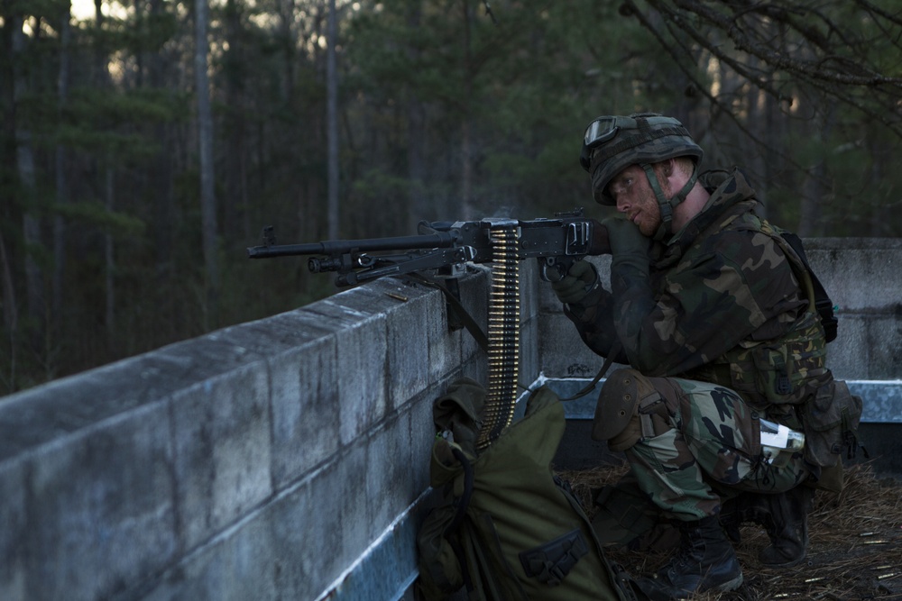 A soldier in camouflage uniform is positioned behind a concrete wall barrier, aiming a belt-fed machine gun in what appears to be a military training exercise. The soldier is equipped with a helmet, goggles, gloves, and body armor, with an ammunition belt feeding into the weapon. The setting is an outdoor wooded area during low-light conditions, likely dawn or dusk.