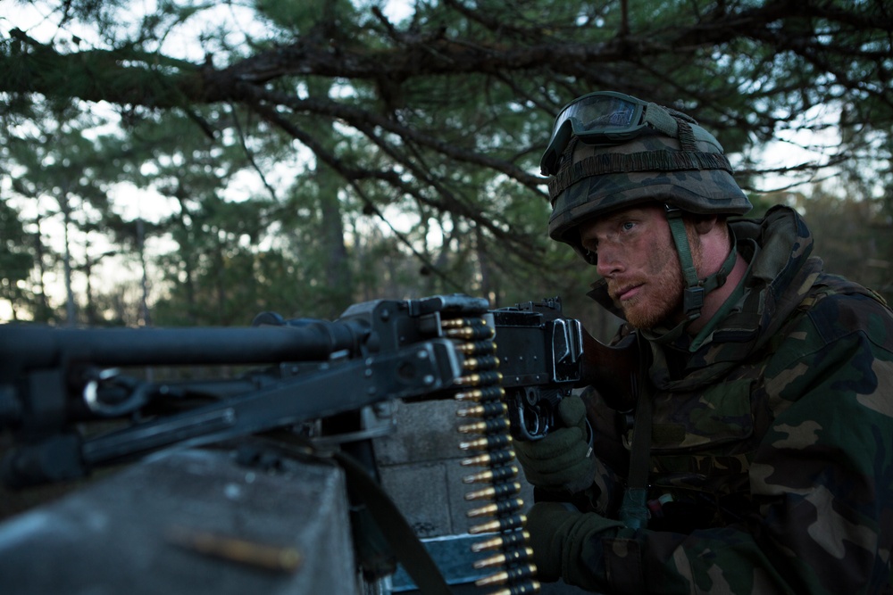 A soldier in woodland camouflage uniform and helmet with mounted goggles is positioned behind a belt-fed machine gun in a forested environment. The soldier appears alert and focused, manning the weapon in what appears to be a training exercise or tactical position. A linked ammunition belt is clearly visible feeding into the weapon.