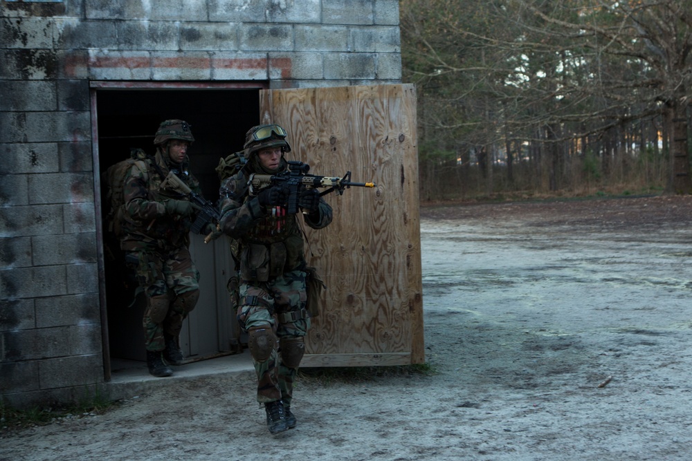 Two military personnel in woodland camouflage uniforms are conducting a building clearance exercise, emerging from a cinder block structure through a wooden door. The lead soldier is advancing with a rifle raised in a ready position, while the second soldier follows from behind. The setting appears to be a military training facility based on the constructed environment and sandy terrain.