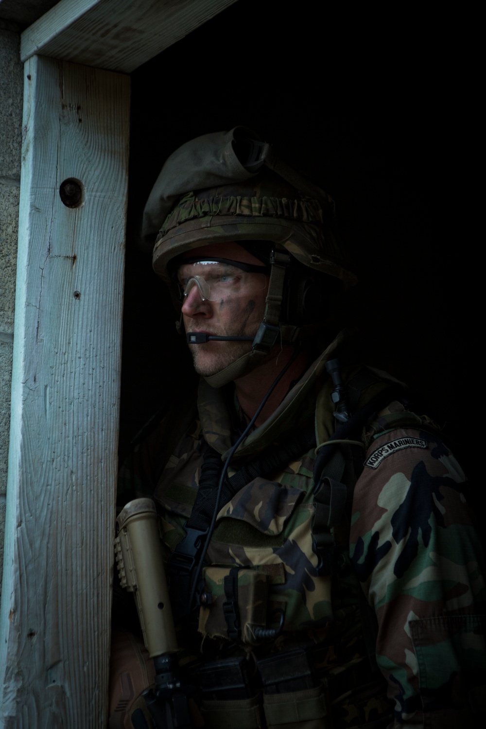 A male soldier in full combat gear is positioned in a doorway, appearing to conduct a tactical entry or surveillance operation. He is wearing Dutch Marine Corps ('Korps Mariniers') insignia on his sleeve, along with a combat helmet, protective eyewear, and a communications headset. The dramatic low-key lighting emphasizes the operational context.