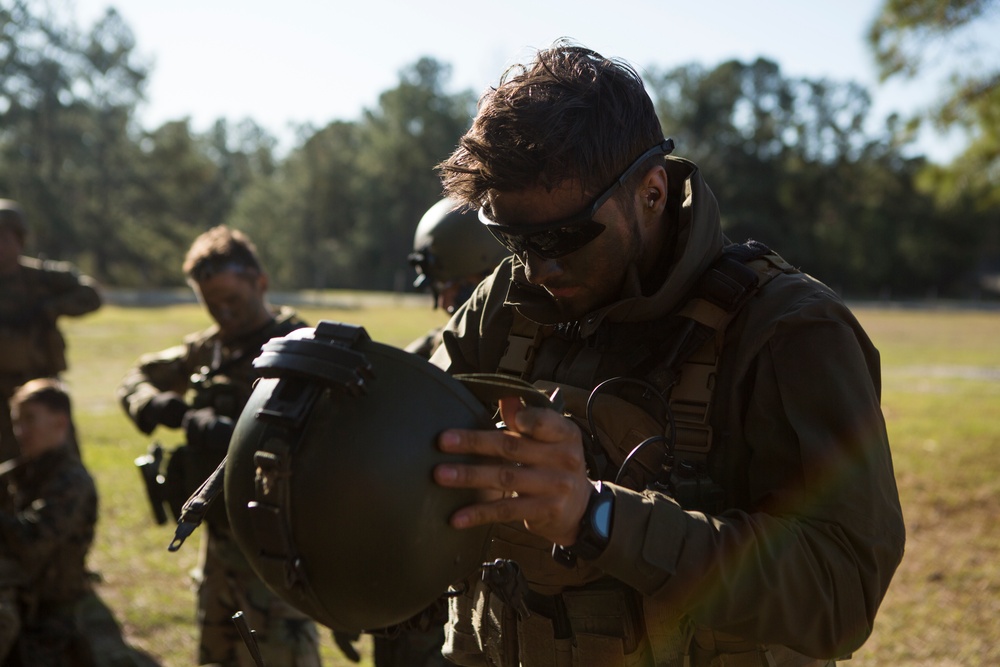 Military personnel in tactical gear are gathered outdoors in what appears to be a training exercise or equipment inspection. The foreground subject is closely examining a ballistic helmet while wearing body armor and tactical sunglasses. Additional personnel are visible in the background performing similar equipment-related tasks.