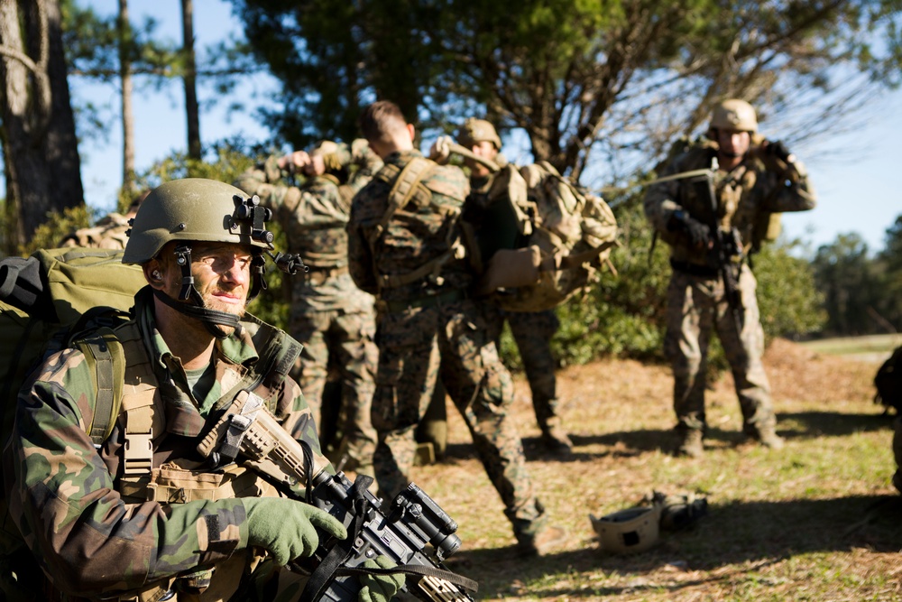 A group of military personnel in camouflage uniforms are gathered in an outdoor wooded area, appearing to be during a training exercise or field operation. The foreground soldier is equipped with a helmet mounted with a night vision device, body armor, tactical gloves, and an assault rifle. Background soldiers appear to be assisting each other with gear and equipment adjustments.