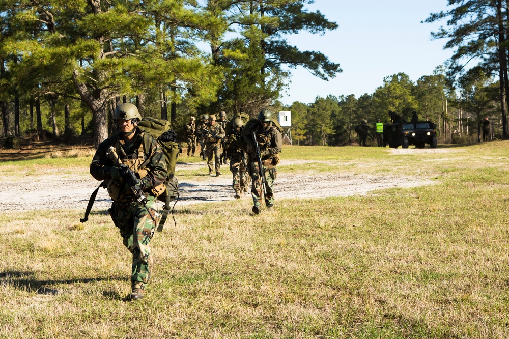 A column of military personnel in full combat gear marches along a sandy dirt road through a pine tree-lined training area. The soldiers are carrying weapons and large rucksacks, suggesting a ruck march or field exercise. A military Humvee and personnel in high-visibility vests are visible in the background, indicating an organized training event.