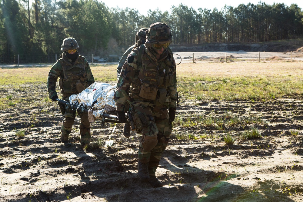 Three military personnel in camouflage uniforms are carrying a casualty on a litter (stretcher) covered with a reflective emergency thermal blanket across an open, muddy field. The scene appears to be a medical evacuation training exercise conducted in a field environment. The soldiers are wearing full combat gear including helmets, protective goggles, and tactical vests.