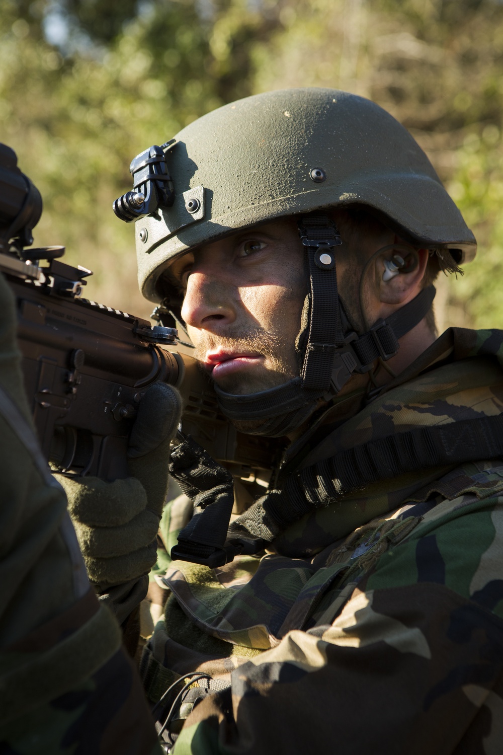 A male soldier in full tactical gear is shown in a close-up portrait, holding a rifle in a ready position. He is wearing woodland camouflage BDUs, a PASGT-style ballistic helmet with a night-vision device mount, and tactical gloves. The outdoor setting features blurred green vegetation in the background, suggesting a field training or operational environment.