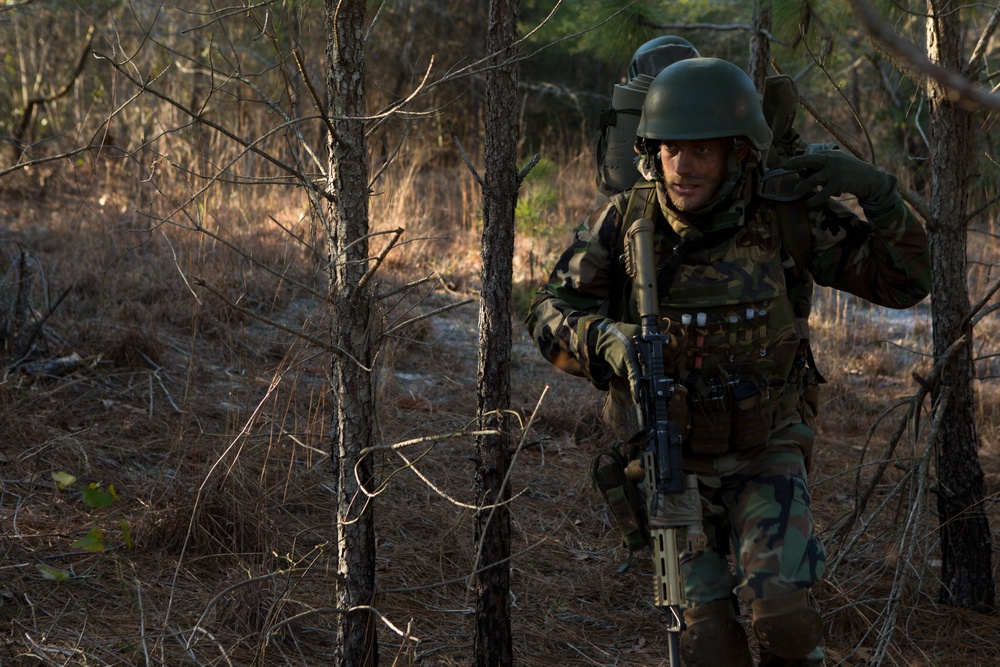 A soldier in full combat gear moves through a sparse woodland environment. The individual is equipped with camouflage uniform, helmet, tactical vest, backpack, and carries an assault rifle. The setting appears to be a training exercise or field operation in a dry, deciduous forest.