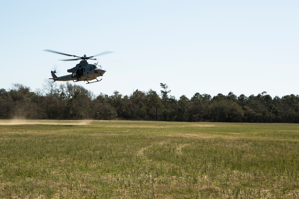 A military helicopter, appearing to be a Bell UH-1Y Venom or similar utility helicopter in gray/olive drab livery, is hovering or taking off at low altitude over an open grassy field. Rotor wash is visibly kicking up dust and debris to the left of the aircraft. The setting appears to be a rural training area bordered by a treeline.