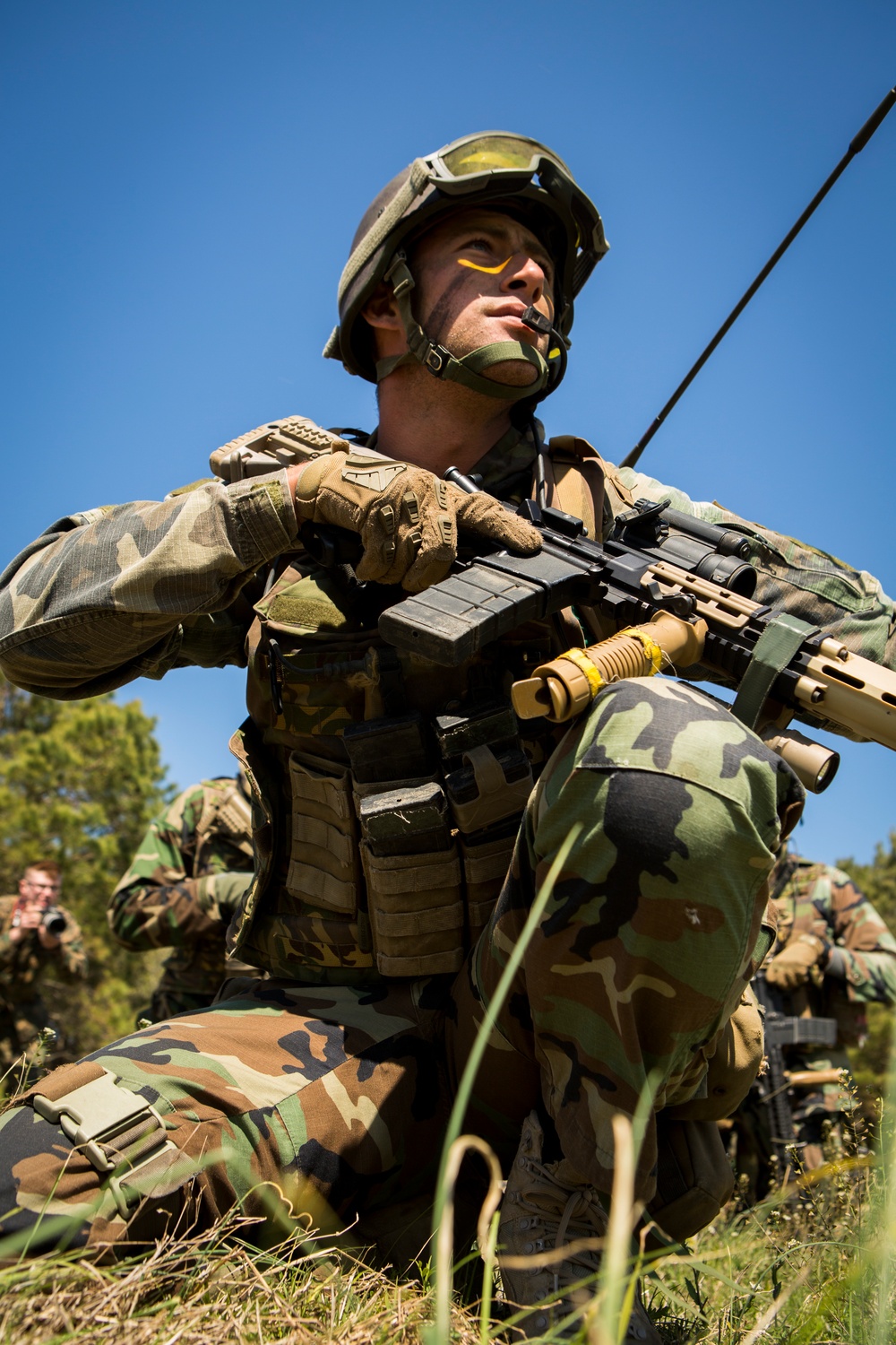 A soldier in woodland camouflage BDUs is kneeling in a field position, holding an assault rifle with a tan/FDE finish and looking upward. The soldier is equipped with full tactical gear including a MOLLE plate carrier, ballistic helmet with goggles, tactical gloves, and face paint in yellow and green. Additional soldiers in similar uniforms are visible in the background, suggesting a training exercise or field operation.