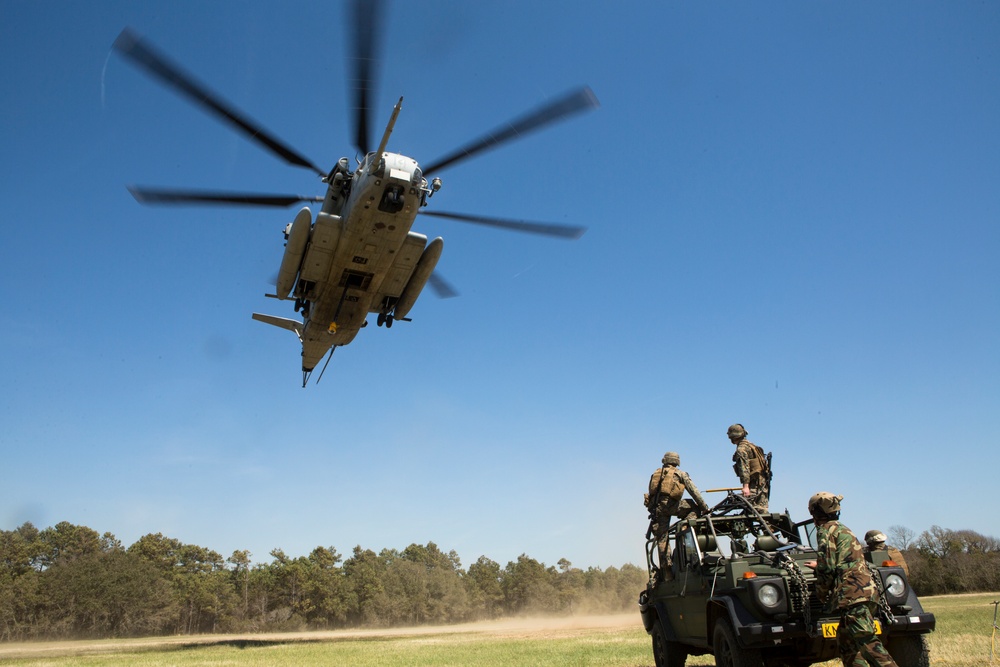 A large military helicopter, appearing to be a CH-53E Super Stallion, hovers low over an open grassy field during what appears to be a military training exercise or operation. Several soldiers in camouflage uniforms are positioned on or around a military light utility vehicle, observing or coordinating with the helicopter. The scene takes place in a clearing surrounded by trees under clear blue skies.