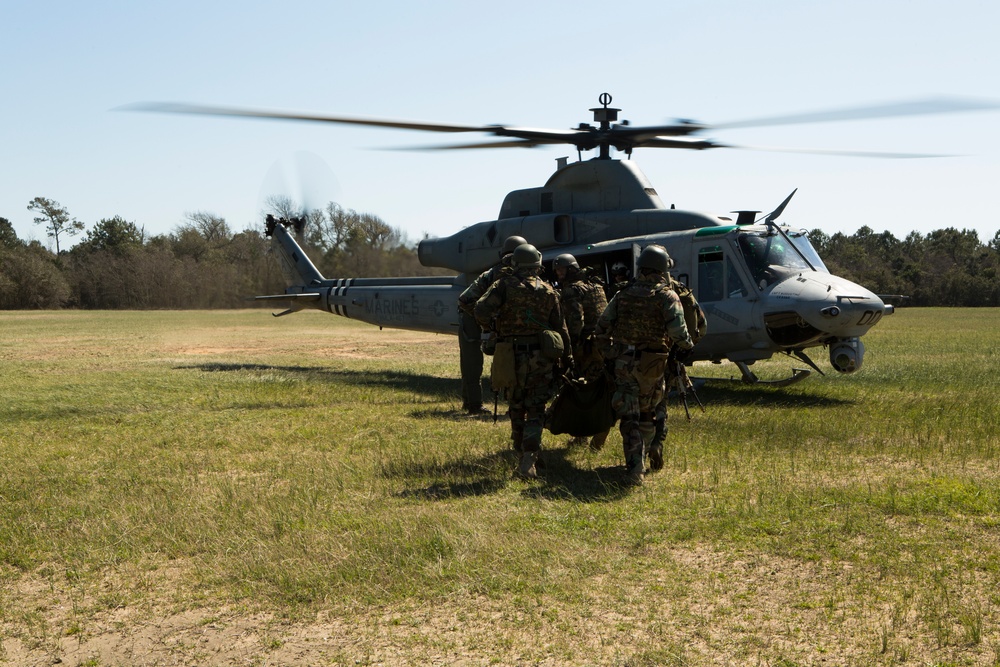 A group of Marines in full combat gear are loading or boarding a U.S. Marines UH-1Y Venom helicopter on an open grassy field. Several personnel appear to be carrying a litter or stretcher, suggesting a medical evacuation (MEDEVAC) or casualty extraction exercise. The helicopter's rotors are spinning, indicating it is either landing or preparing for immediate takeoff.