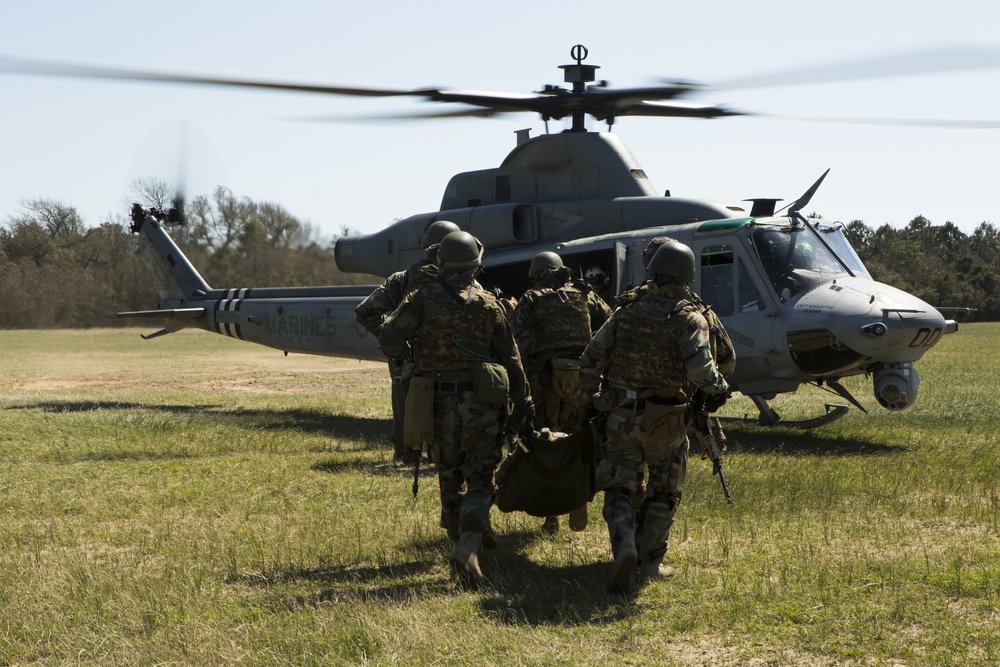 A group of military personnel in camouflage uniforms are carrying a litter (stretcher) toward a U.S. Marines helicopter on a grassy field. The helicopter, identifiable by 'MARINES' markings on the fuselage, appears to be a UH-1Y Venom with rotors spinning. This appears to be a medical evacuation (MEDEVAC) exercise or operation.
