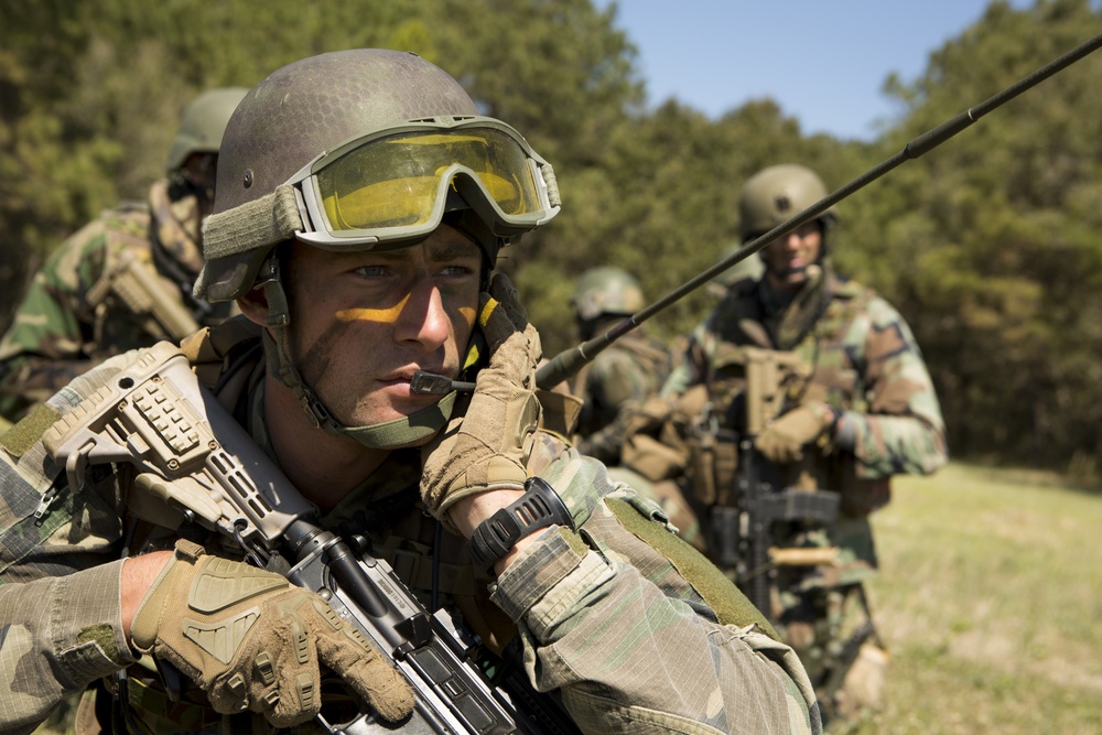A group of soldiers in full combat gear are positioned in an outdoor field environment. The foreground soldier is using a radio communication device while holding a rifle, with face camouflage paint applied. Additional soldiers are visible in the background, suggesting a coordinated military field exercise or operation.