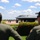 Two military personnel in olive/sage green flight suits and black caps are observing a B-2 Spirit stealth bomber on a tarmac. The aircraft, marked with 'WM' tail code indicating assignment to Whiteman Air Force Base, is surrounded by ground crew personnel. The scene appears to be at a military airfield in a tropical or subtropical environment.