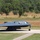 A B-2 Spirit stealth bomber is taxiing on a runway at what appears to be a military airfield. The aircraft displays its distinctive flying-wing design with a dark gray low-observable coating. Trees and dry grassland are visible in the background, suggesting a warm, possibly semi-arid environment.