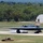 A B-2 Spirit stealth bomber is landing or taking off on a military airfield runway, with visible smoke/exhaust from its engines and landing gear deployed. The aircraft's distinctive flying-wing design is clearly recognizable against the flat runway surface. A Quonset-style hangar structure and tree line are visible in the background.