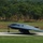A B-2 Spirit stealth bomber is taxiing on a runway, likely at a military airbase. The aircraft's distinctive flying-wing design is clearly visible, with its flat, blended profile and dark gray stealth coating. Green tree-lined terrain is visible in the background, suggesting a temperate environment.