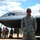 A U.S. Air Force airman in ABU (Airman Battle Uniform) stands at attention in front of a B-2 Spirit stealth bomber on a flight line. The aircraft bears the name 'Spirit of Indiana' visible on its fuselage. Several other personnel are gathered in the background near the aircraft.