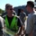 A man wearing a bright neon yellow/green safety vest and sunglasses appears to be speaking with military personnel in uniform at what looks like a military installation or airfield. The central figure is engaged in conversation with a younger man in a light-colored t-shirt, while several other uniformed service members are visible in the background. The setting appears to be an outdoor area with a building and trees visible behind the group.