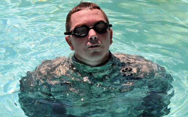 A person is standing in a swimming pool while wearing military camouflage uniform (ACU pattern) and black swimming goggles. The individual appears to be submerged to approximately chest level in clear, blue-green pool water. This appears to be a military water survival or combat swimming training exercise.