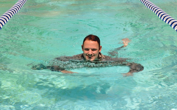 A young man with red/auburn hair is swimming in a clearly marked lap pool with blue and white lane dividers. He is smiling at the camera with his head above water, arms extended outward in what appears to be a breaststroke position. The pool water is clear and turquoise, suggesting an outdoor or well-lit facility.