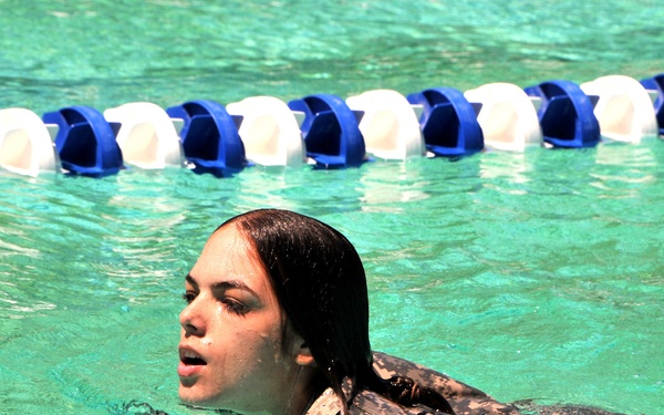 A swimmer is photographed in an outdoor or indoor competition pool, positioned between two lane dividers made of alternating blue and white floats. The swimmer, with wet dark hair, appears to be resting or recovering after a swim, with their head just above the water surface. The pool water has a distinctive turquoise-green color.